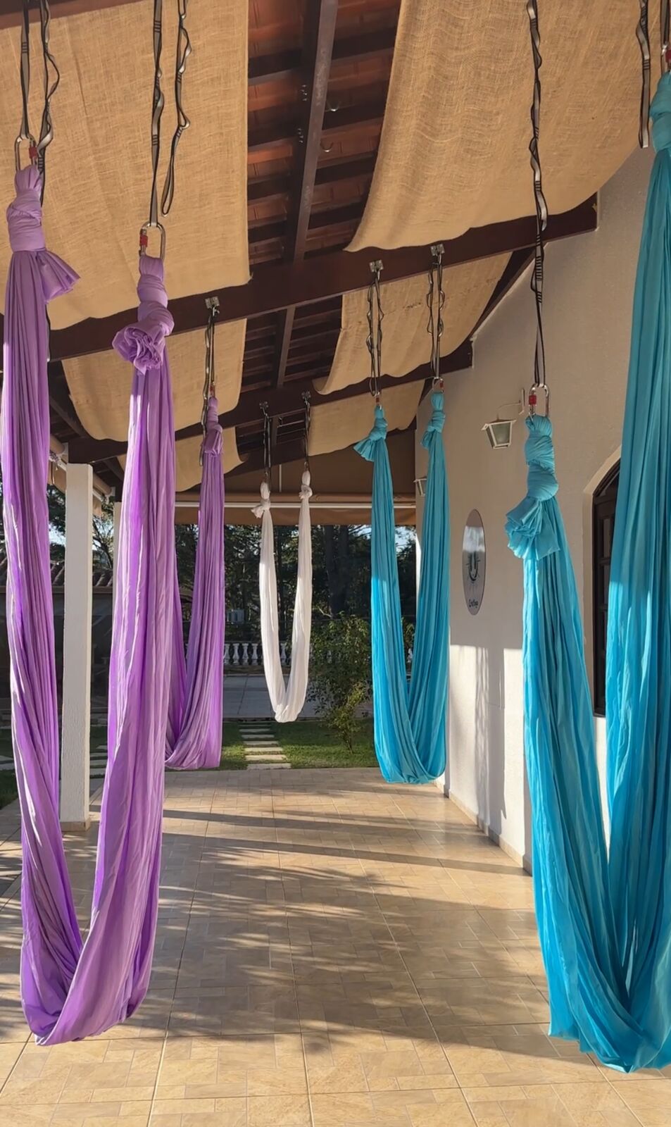 Vista ampla do espaço coberto do studio, com tecidos de Aerial Yoga e jardim ao fundo